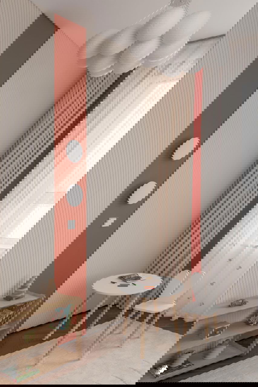 Детская зона с небольшим столиком и стулом в виде зайчика, вертикальной красной полосой на стене и светильником из круглых шаров — мягкая и уютная композиция.