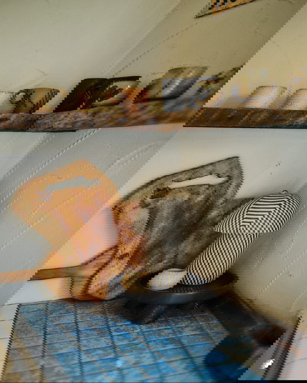 Угловая полка с керамическими кружками, декоративная лампа в полоску и разделочная доска на фоне шероховатой штукатурки — рустикальные детали интерьера.