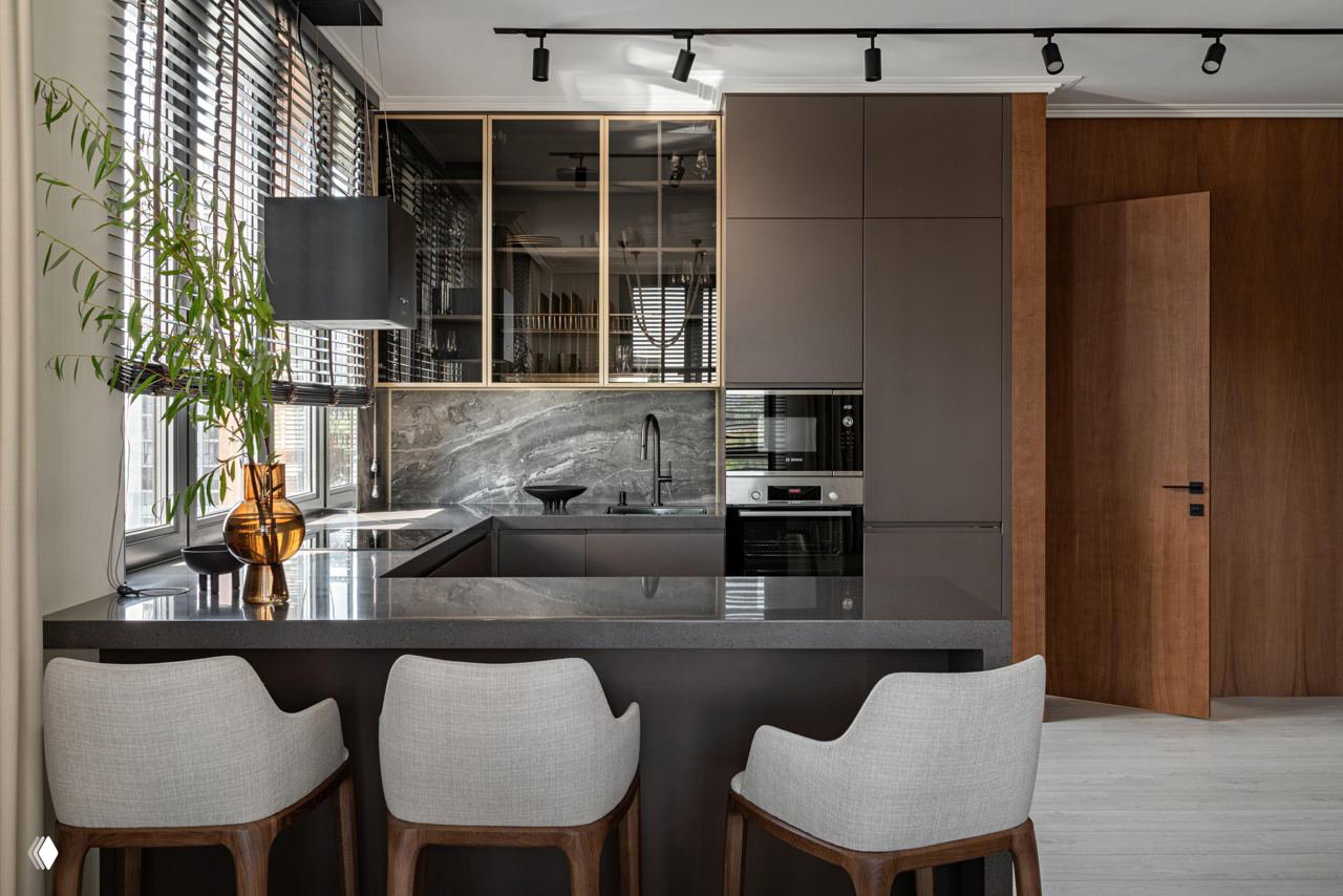 Кухня-гостиная с барной стойкой и тёмной столешницей, стеклянные верхние шкафы, мраморный фартук и три барных стула в современном оформлении.