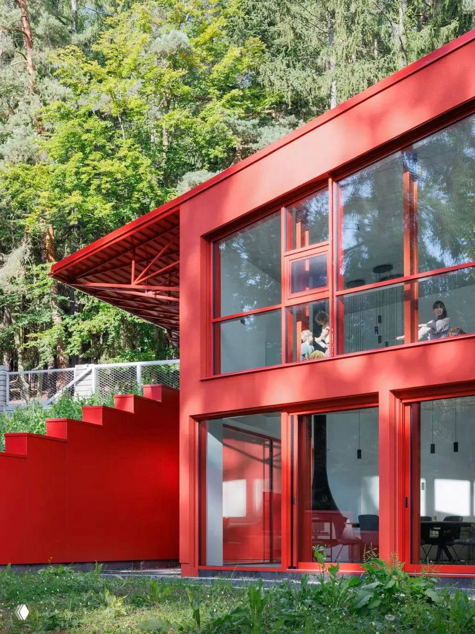 Крупный план фасада с большими панорамными окнами: красные металлические рамы и ступенчатая кровля на фоне зеленых крон высоких деревьев чешского леса.