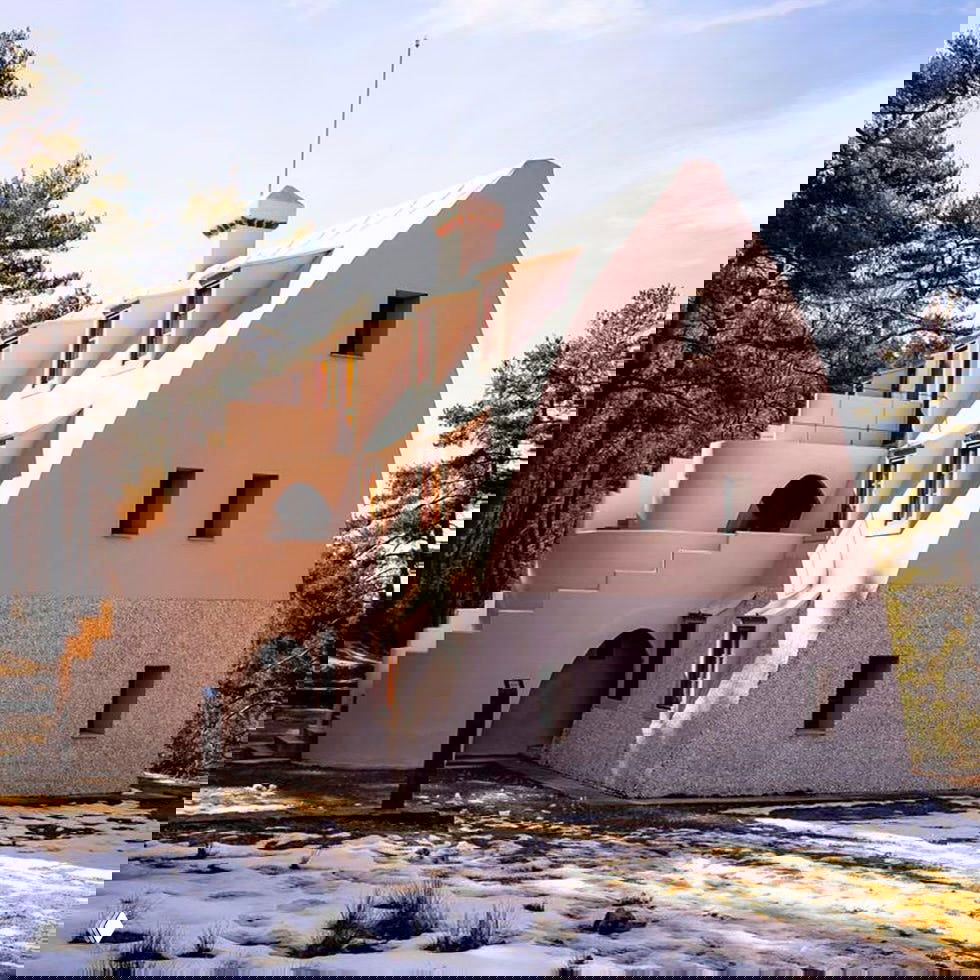 Фасад дома в розовом оттенке: ступенчатая наружная лестница, окна с деревянными рамами и лесной пейзаж вокруг.