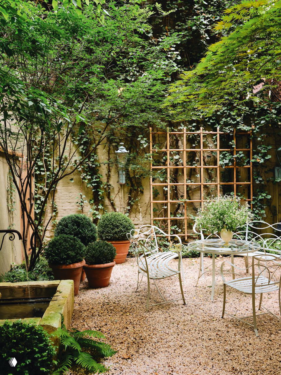 Уютный закрытый садик во внутреннем дворе: кованая садовая мебель, горшки с самшитом, решетка для вьющихся растений и зелёная живая растительность.