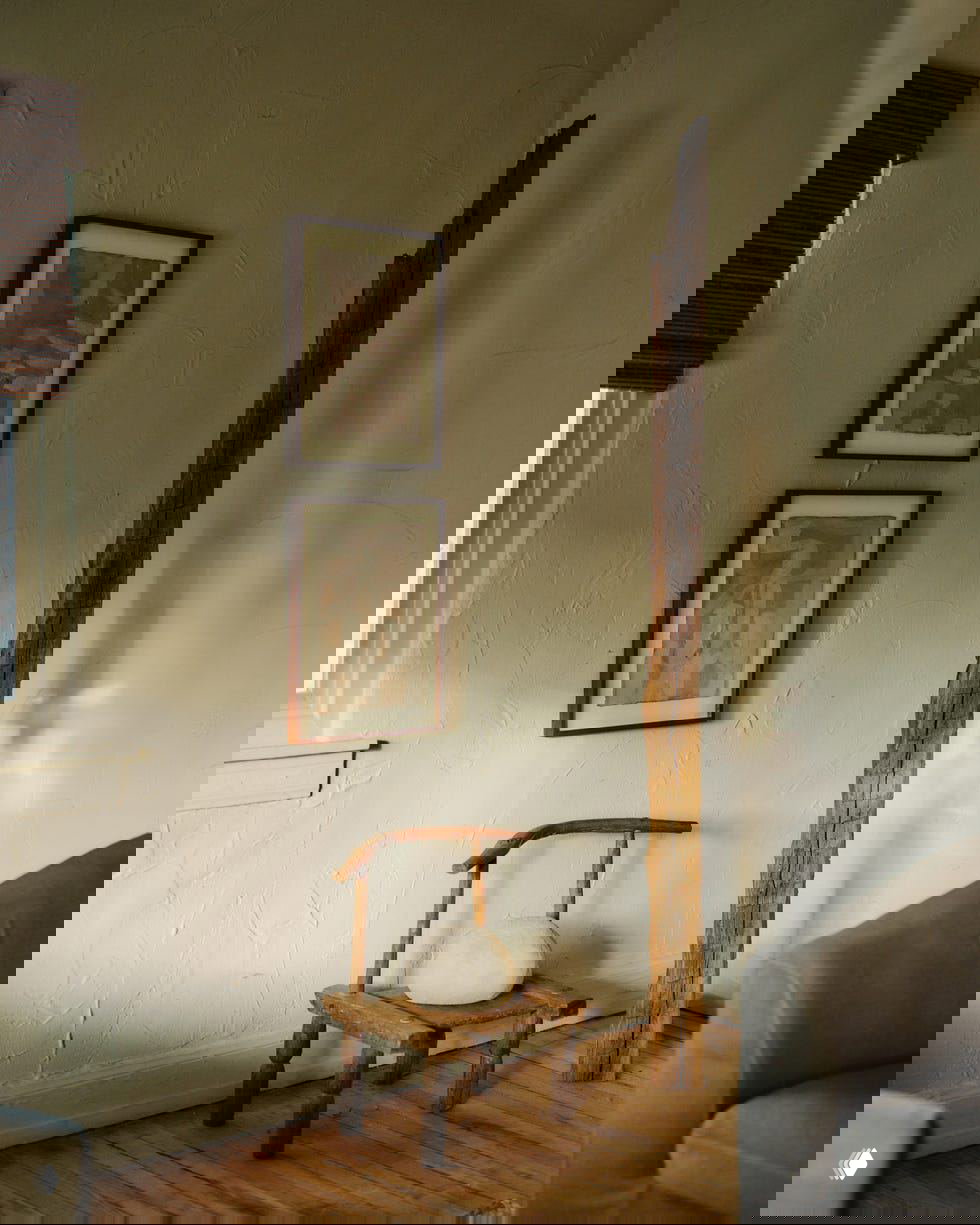 Небольшой деревянный стул и резной высокий столб у оштукатуренной стены с двумя рамками картин — минималистичный уголок с текстурой и деревом.