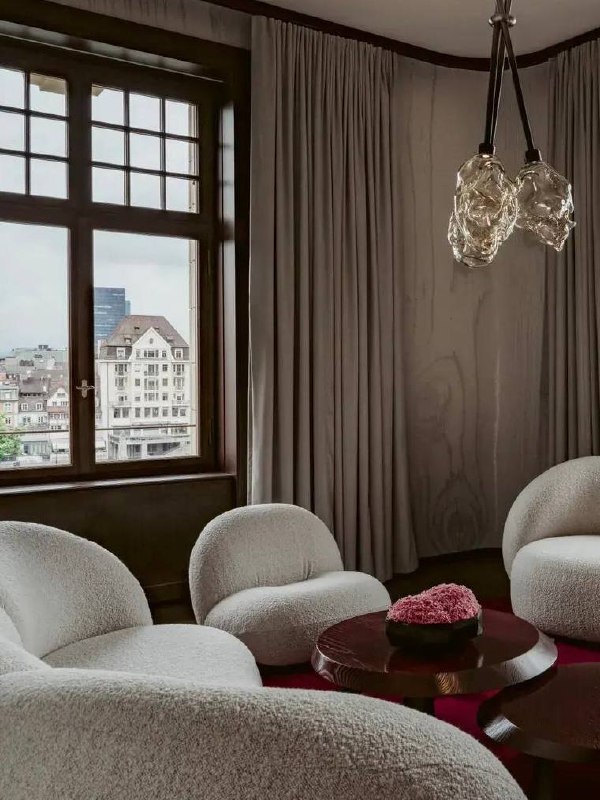 Уютная гостиная люкса с белыми креслами boucle, низкими столиками и видом на город через большое деревянное окно — спокойная зона для отдыха в новом крыле.