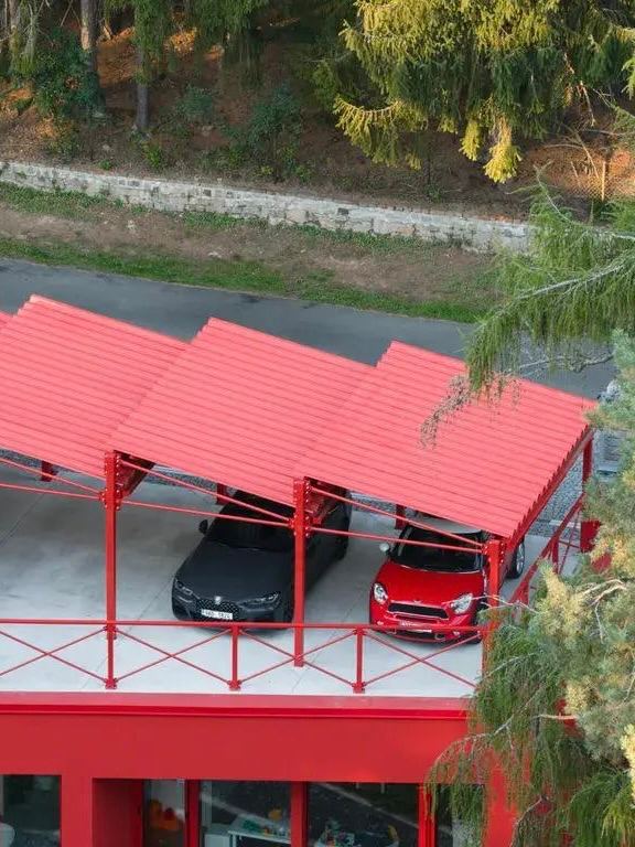 Красная навесная парковка рядом с домом: металлическая конструкция и крыша насыщенного красного цвета, два автомобиля под навесом, окружение леса.