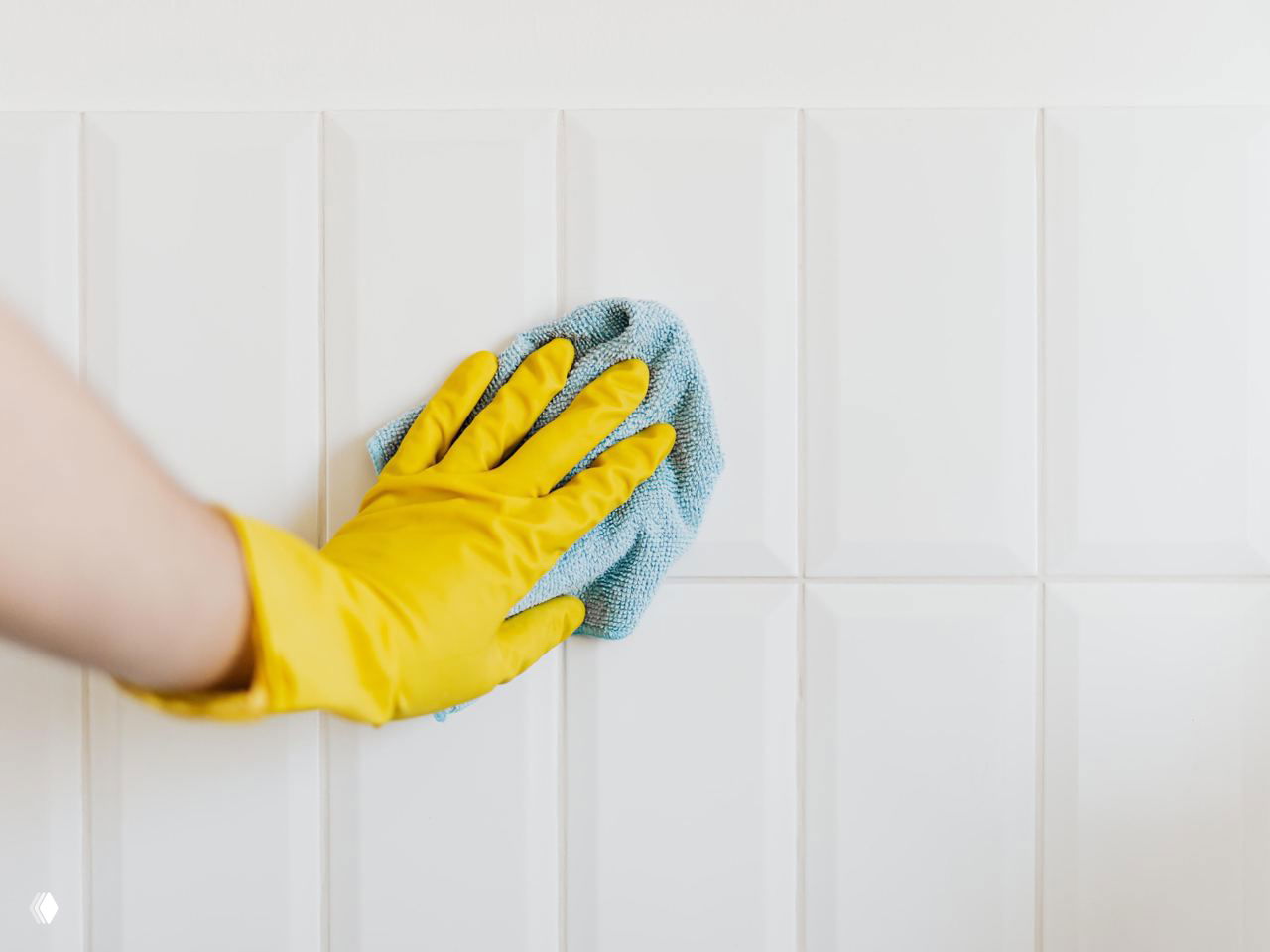 Рука в жёлтой резиновой перчатке протирает белую кафельную плитку мягкой тряпкой — уборка и подготовка поверхности.
