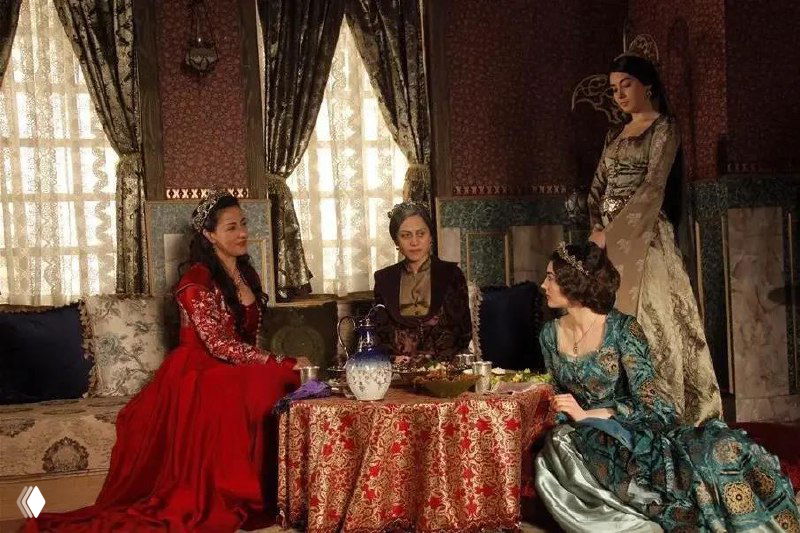 Группа женщин в роскошных платьях сидит вокруг низкого стола с кувшином и яствами, на заднем плане узорчатые ткани и драпировки.