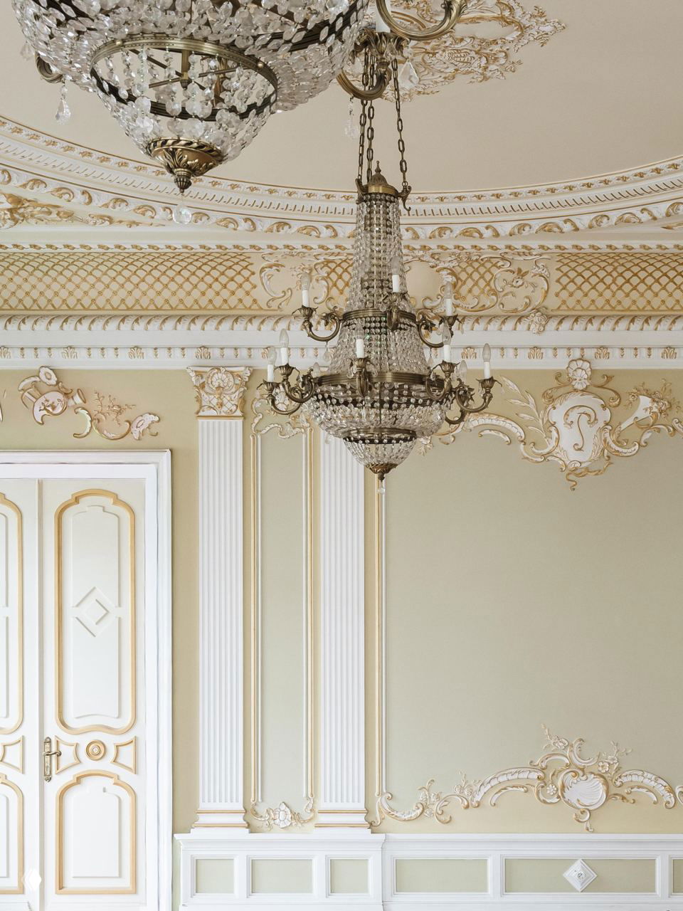 Зал с богато декорированным потолком и хрустальной люстрой: сложная лепнина, профилированные карнизы и массивная подвесная люстра с кристаллами на фоне пастельных орнаментов.