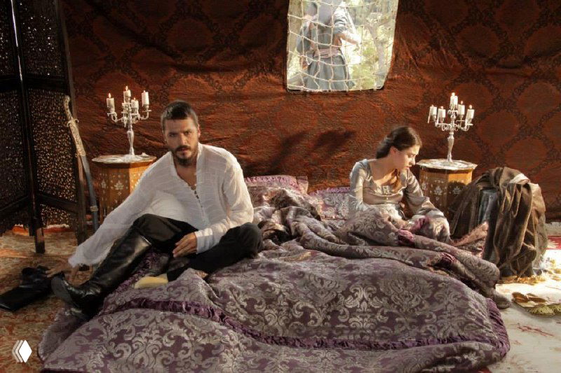 Комната с большим покрывалом и алюминиевыми подсвечниками по бокам, на кровати мужчина и женщина в костюмах — декоративные элементы интерьера в кадре.