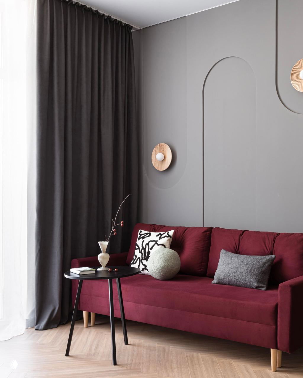 Бордовый диван у окна, тёмно-серые шторы, декоративная серая стеновая панель с арочными элементами и компактный круглый кофейный столик.