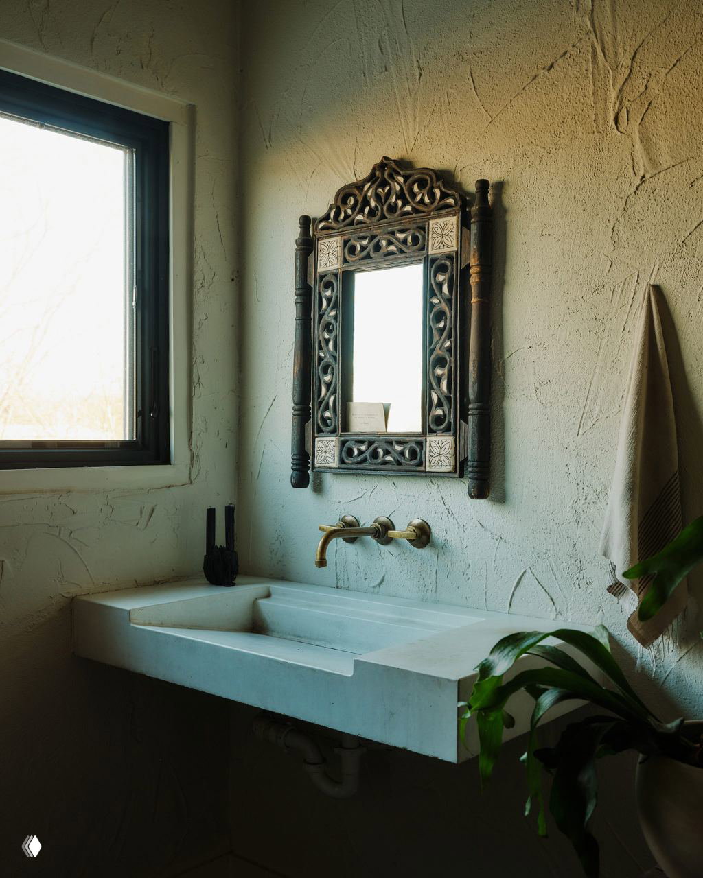 Раковина из светлого камня с бронзовым смесителем и резным зеркалом на текстурированной штукатурке; растительный акцент у окна.