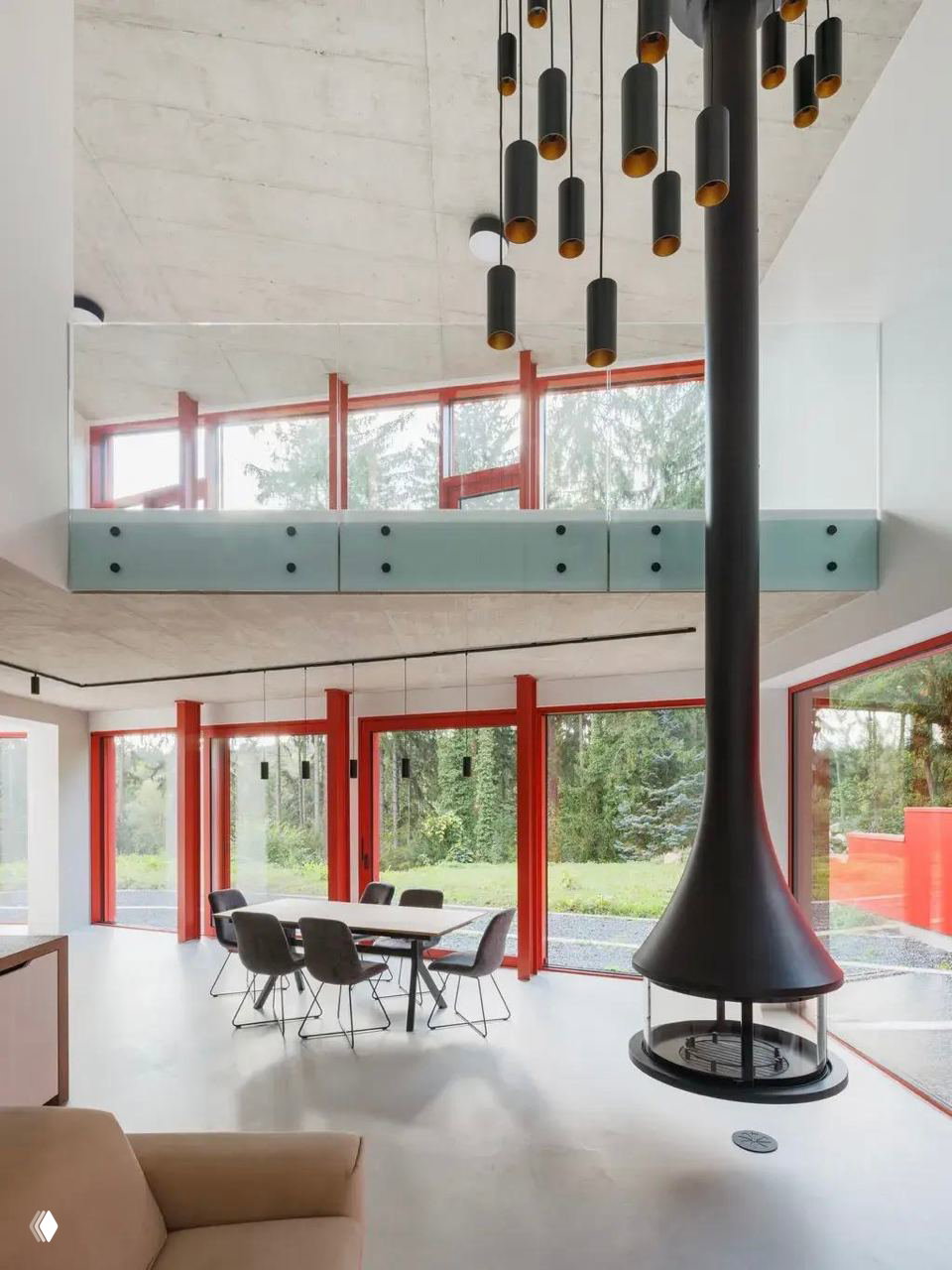 Интерьер первого этажа с открытой планировкой: белые стены, красные оконные рамы и центральный подвесной камин, через окна видна лесная зелень снаружи.