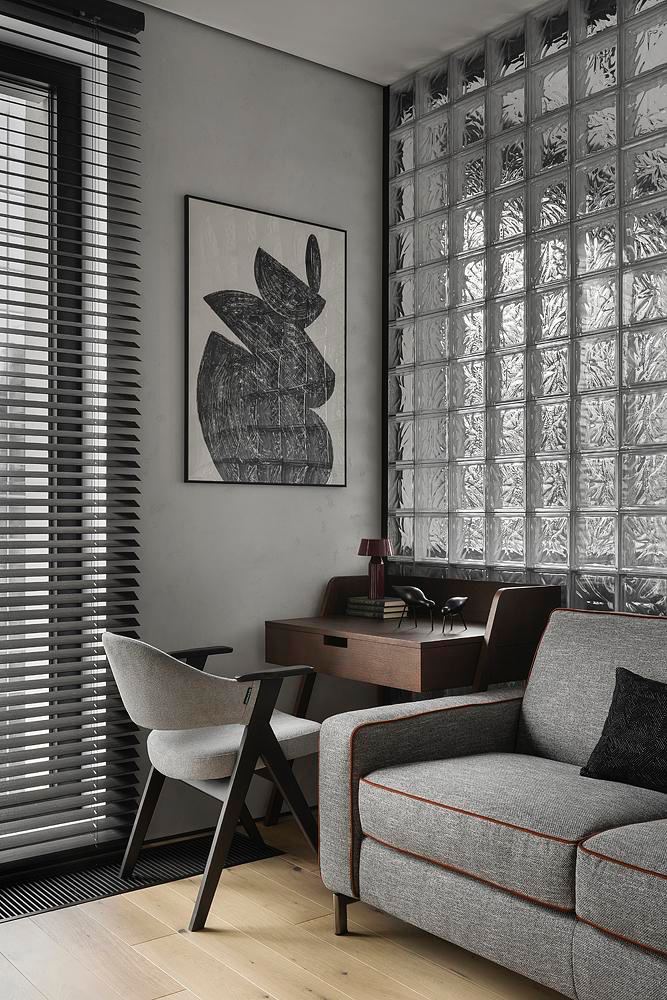 Гостиная с большой стеной из стеклоблоков, диваном и рабочим столом у окна с жалюзи — игра света и прозрачных поверхностей в интерьере.