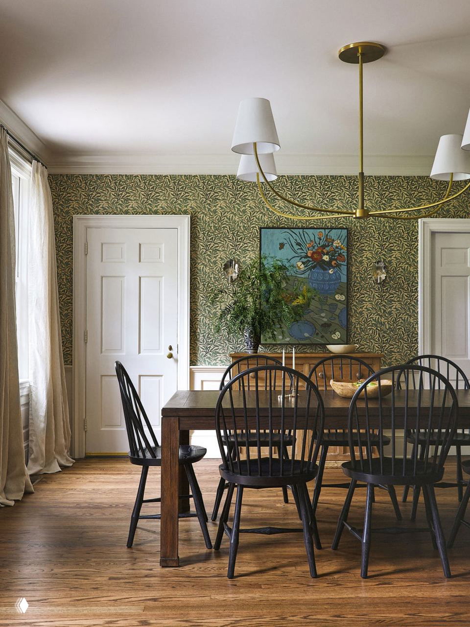 Столовая с длинным деревянным столом и набором чёрных шпиндл-стульев, узорная обивка стен и большая люстра — сочетание классики и уютной семейной обстановки.
