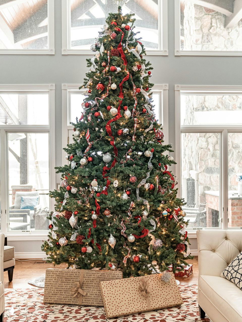 Большая классическая рождественская ёлка в светлой гостиной, украшенная лентами, шарами и огоньками, с подарками у основания.