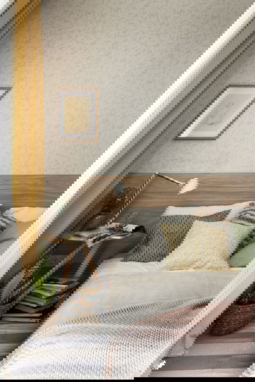 Спальная зона с деревянным изголовьем, несколькими декоративными подушками и ночным столиком с лампой и книгами в маленькой квартире.