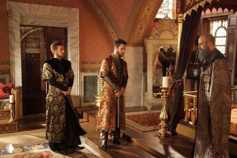 Интерьерный кадр: сцена со сложными текстильными орнаментами и узорчатой отделкой стен, видны декоративные ковры и драпировки.