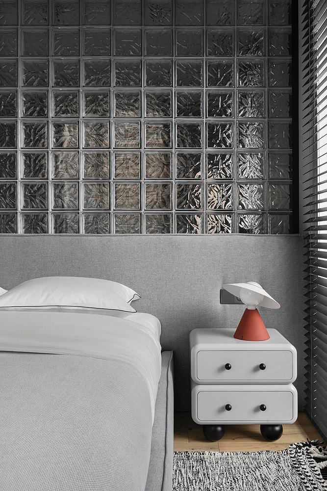 Спальня с изголовьем на фоне стены из стеклоблоков, прикроватная тумба и текстиль в спокойных серых тонах, минималистичный декор.