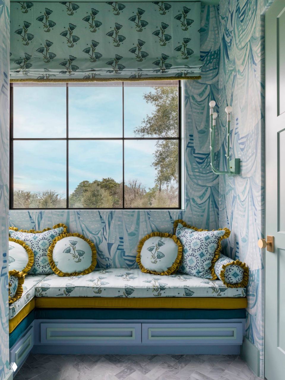 Небольшая уютная ниша у окна: встроенное мягкое сиденье с декоративными подушками, светлые обои с растительным узором и вид на участок ранчо.