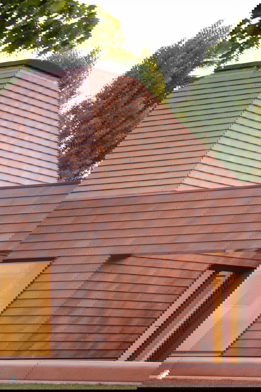 Крупный план терракотовой облицовки и пирамидальной кровли детского сада AACM, видна текстура плитки и мягкий тёплый свет заката на поверхности