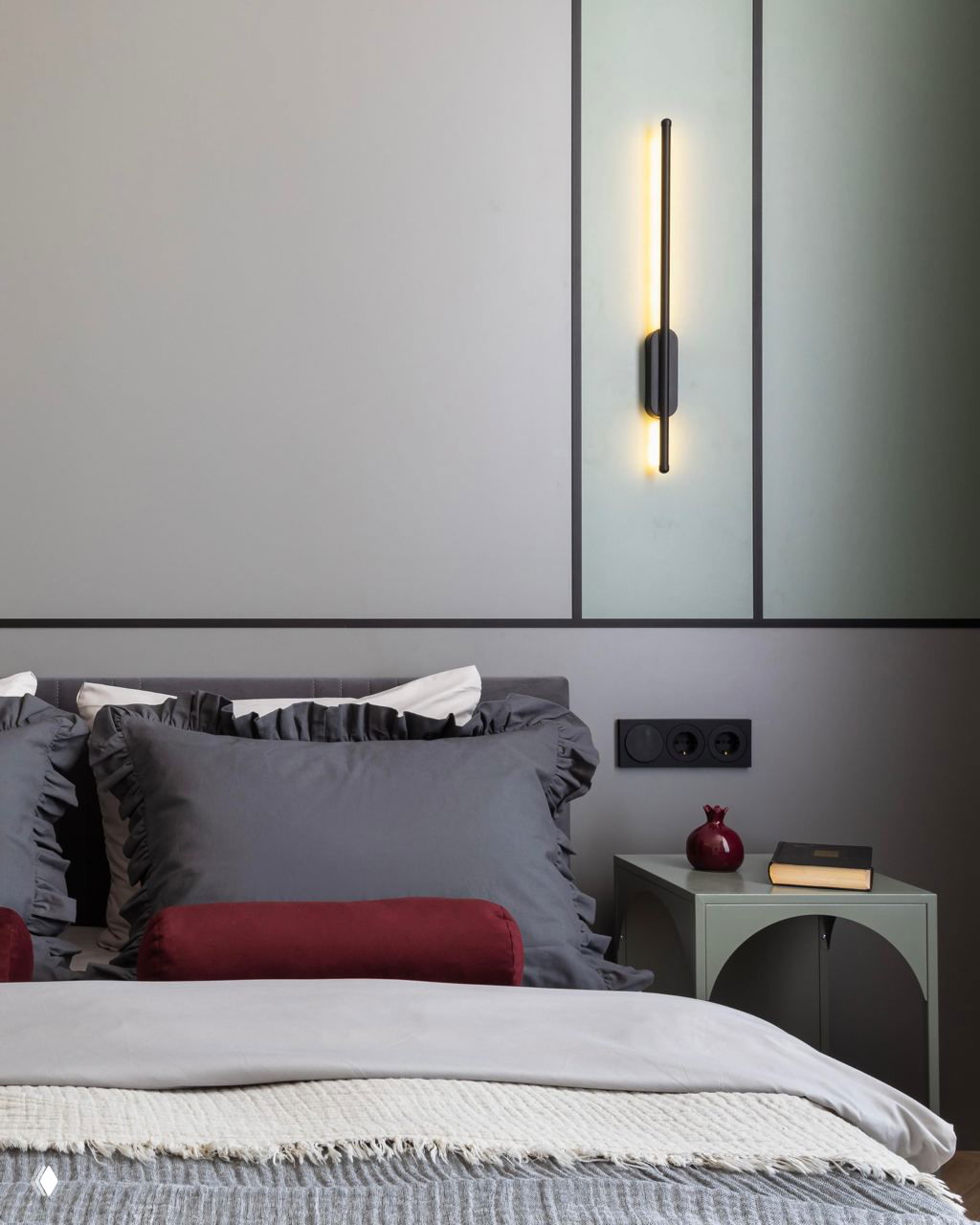 Спальня с серой текстильной отделкой: подушки, валики на кровати, вертикальный тонкий светильник на стене и прикроватная тумба пастельного тона.