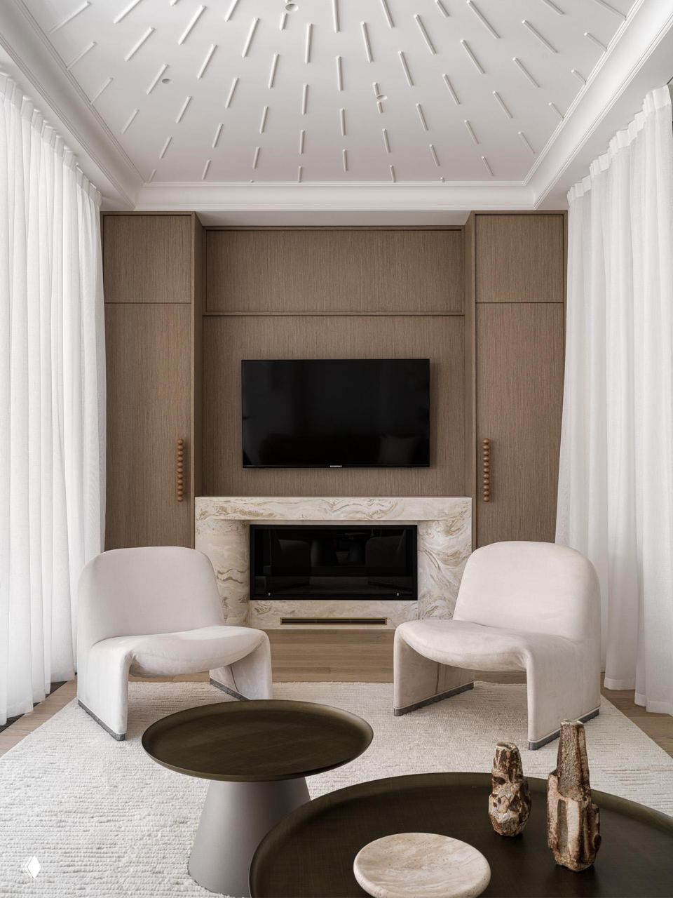 Минималистичная гостиная с телевизором над мраморным камином, двумя мягкими креслами и круглыми столиками на фоне тонкой отделки стен.