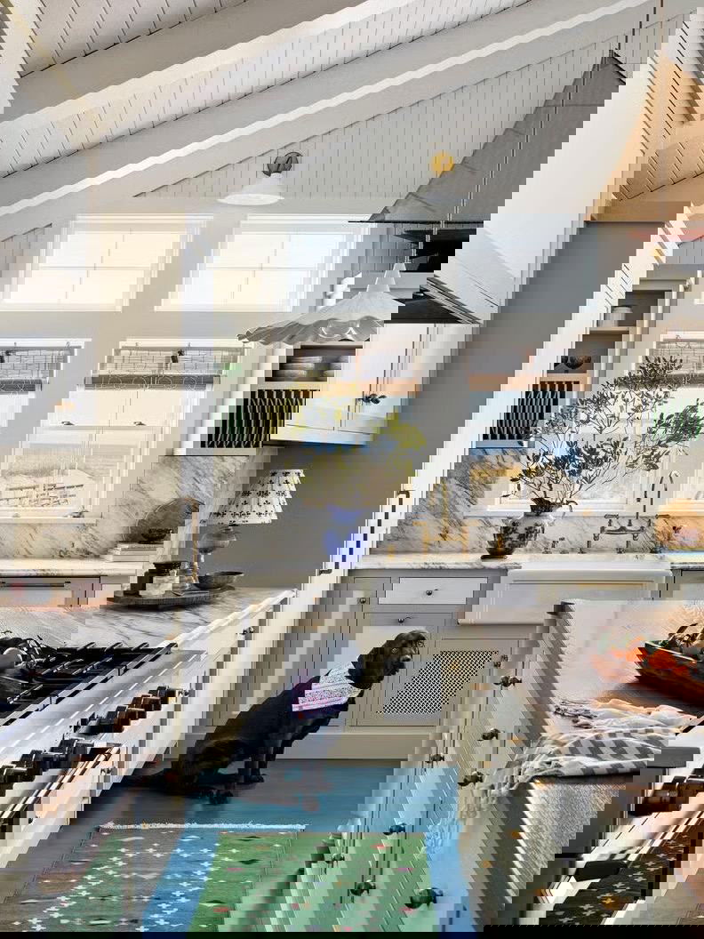 Уютная побережная кухня с бело-голубыми фасадами, ярким синим участком пола и ковриком, у окна видна морская панорама — лёгкий морской стиль