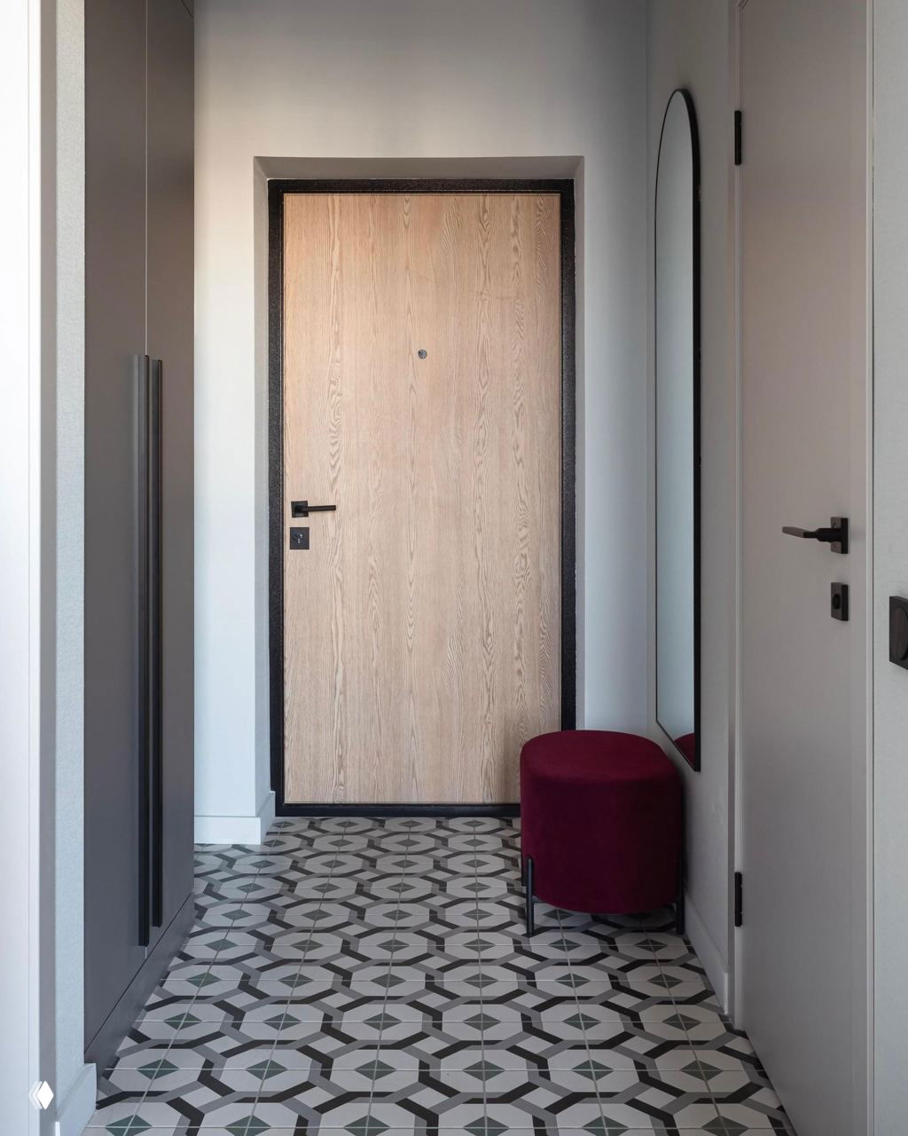 Прихожая с декоративной чёрно‑белой плиткой, светлой деревянной входной дверью, высоким зеркалом и бордовым пуфом у стены для удобства гостей.