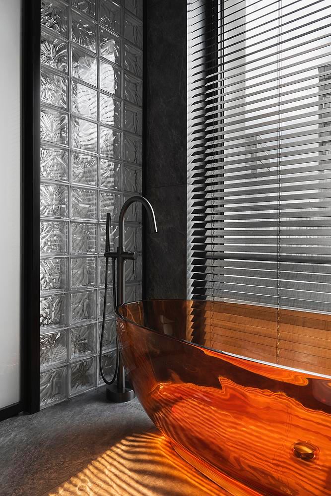 Прозрачная оранжевая ванна на фоне стеклоблоков и тёмных жалюзи — яркий акцент и атмосферная подсветка в компактной ванной комнате.