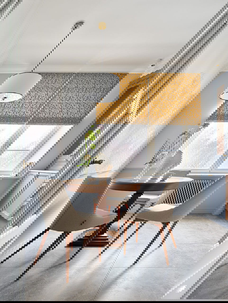 Уютная столовая у окна с круглым деревянным столом, обитыми креслами и яркими римскими шторами на фоне пастельных стен.