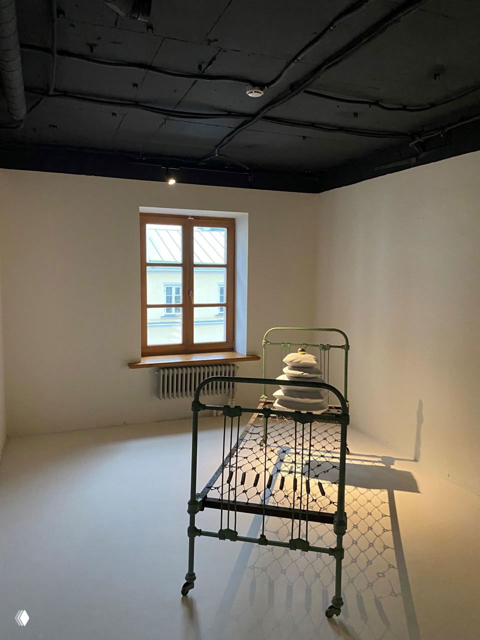 Экспозиция тотальной инсталляции в небольшой белой комнате: металлическая детская кроватка с художественно уложенными предметами и мягкое направленное освещение.