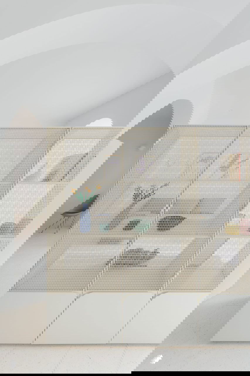 Фрагмент комнаты с полупрозрачной ажурной секцией шкафа на переднем плане; видны вазы, книги и мягкий диван за легкой решеткой в светлом пространстве.