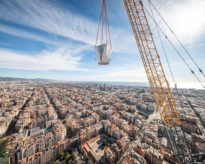 Кран поднимает конструкцию возле Саграда Фамилия: вид сверху на плотную городскую застройку Барселоны и здания вокруг.