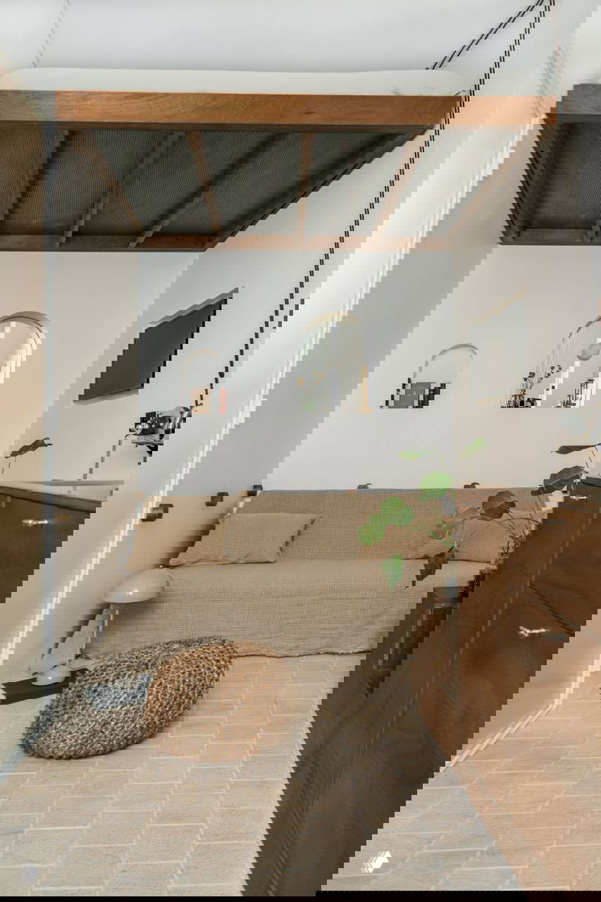 Вид комнаты с поднятой кроватью: диван, плетёный пуф и деревянные фасады справа, растения добавляют живой акцент.