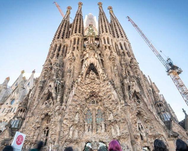 Фрагмент фасада Саграда Фамилия крупным планом с детальной скульптурной резьбой, строительными лесами и кранами у основания портала.