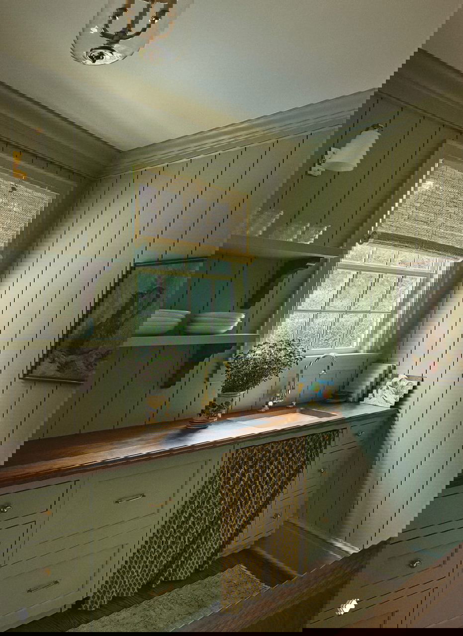 Маленькая утилитарная кухня с вертикальной вагонкой, деревянной столешницей и золотистой фурнитурой крана у окна, приглушённое естественное освещение и уютный вид.