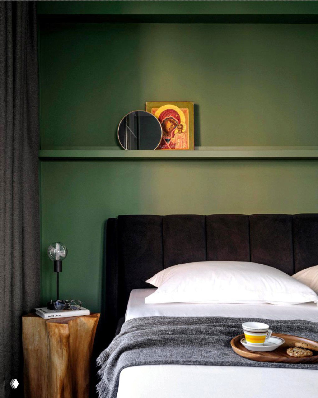 Современная спальня с зелёной стеной и полкой, где стоит малая икона рядом с круглым зеркалом и настольной лампой в интерьере.