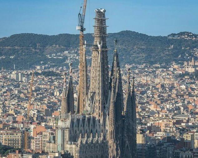 Силуэт Саграда Фамилия с рядами башен и центральной конструкцией в завершающей стадии строительства, вид на город и подъемники.