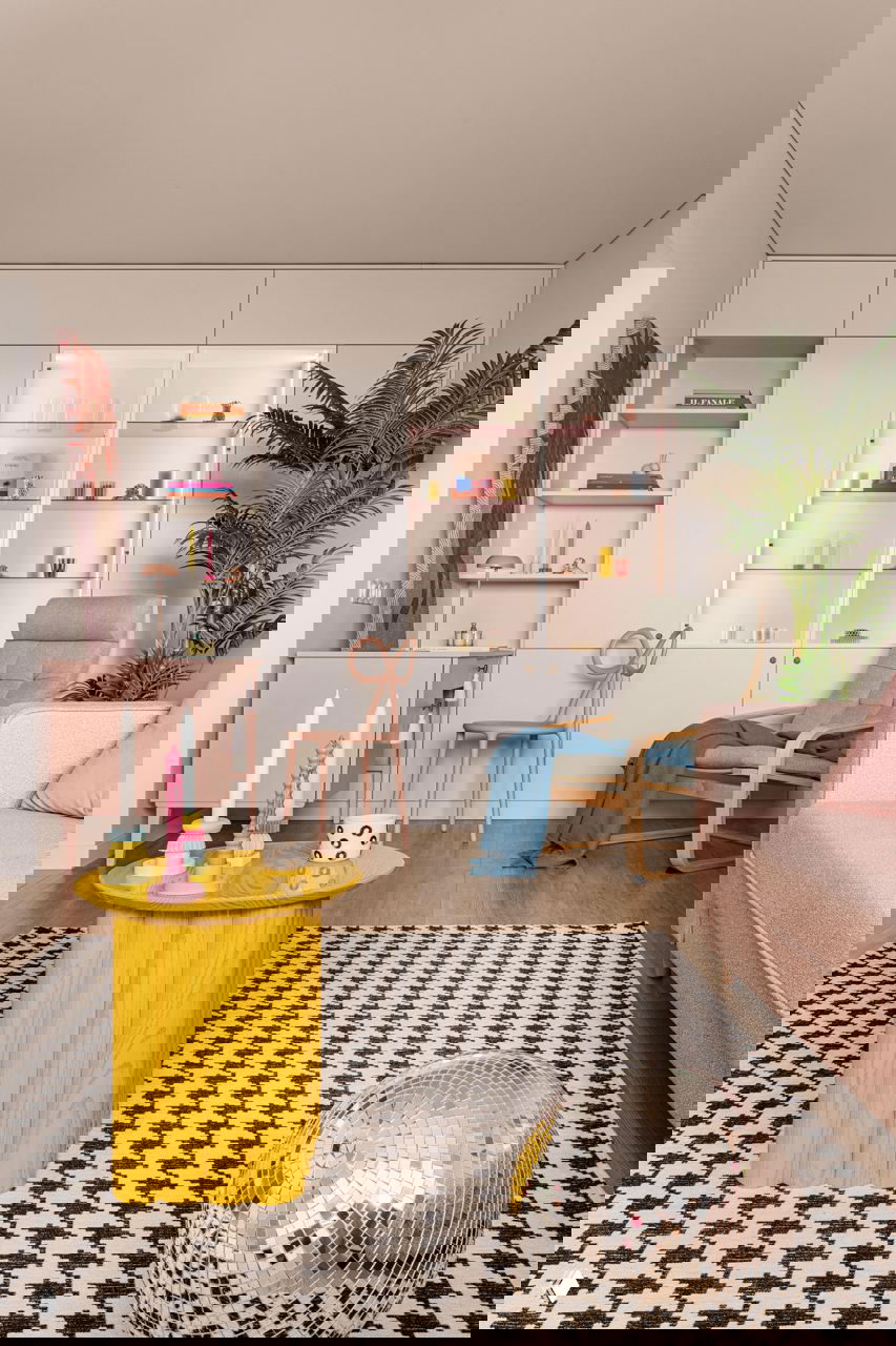 Широкий план гостиной с белыми встроенными шкафами, подсвеченными полками, креслом‑качалкой, розовым рабочим столом, ковром с графическим узором и жёлтым столиком на переднем плане.