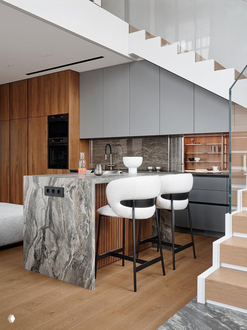 Кухонная зона с островом из натурального камня, барными стульями и деревянными фасадами шкафов под лестницей на антресольном этаже в светлом интерьере.
