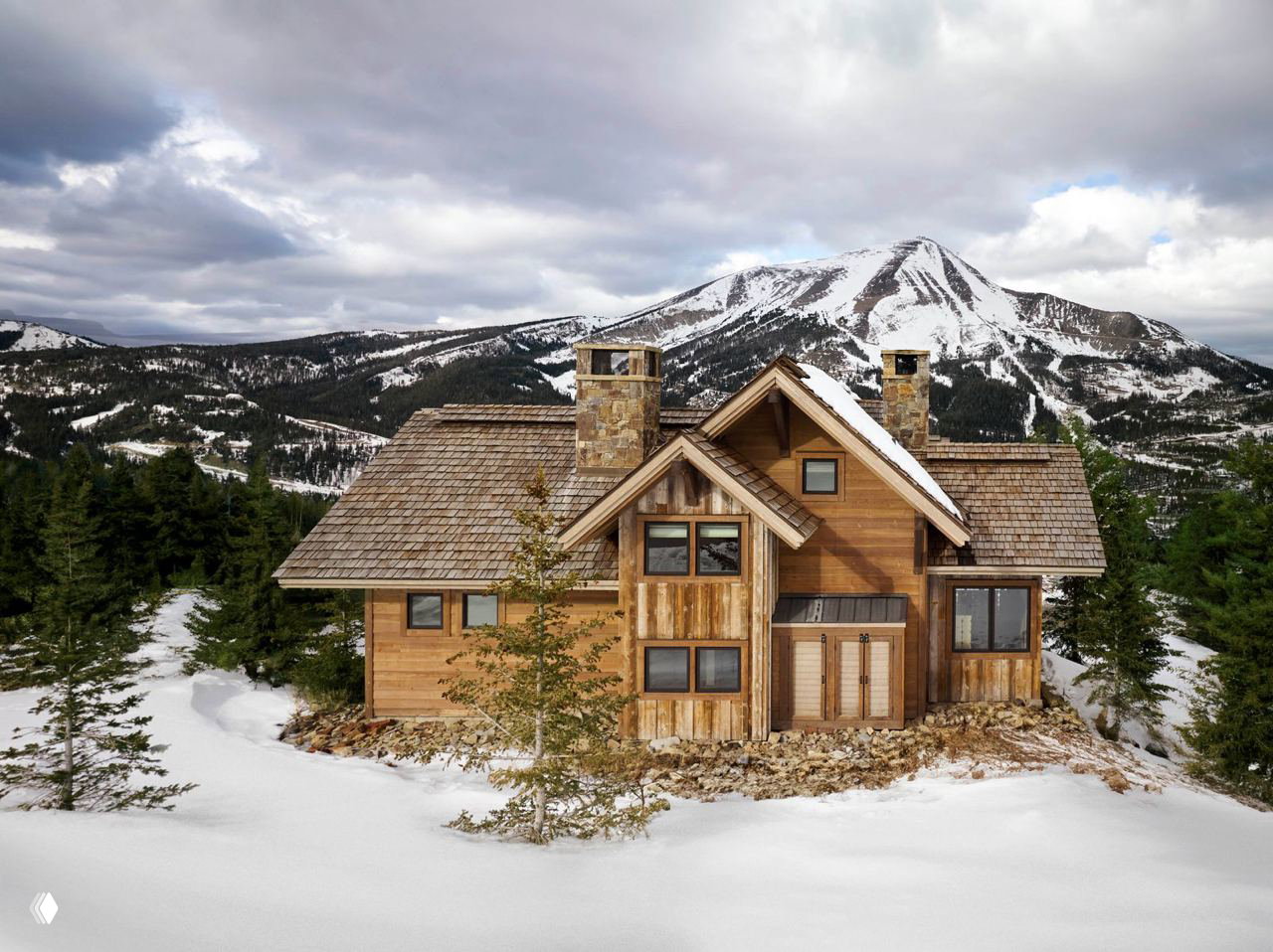 Фасад деревянного шале на заснеженном склоне в Монтане: дом с каминными трубами, деревянной кровлей и видом на горы.