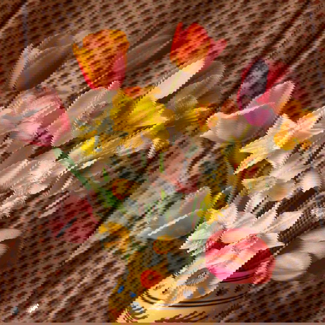 Крупный план букета в керамической вазе: тюльпаны и нарциссы тёплых тонов на фоне узорчатой ткани, интерьерная композиция.