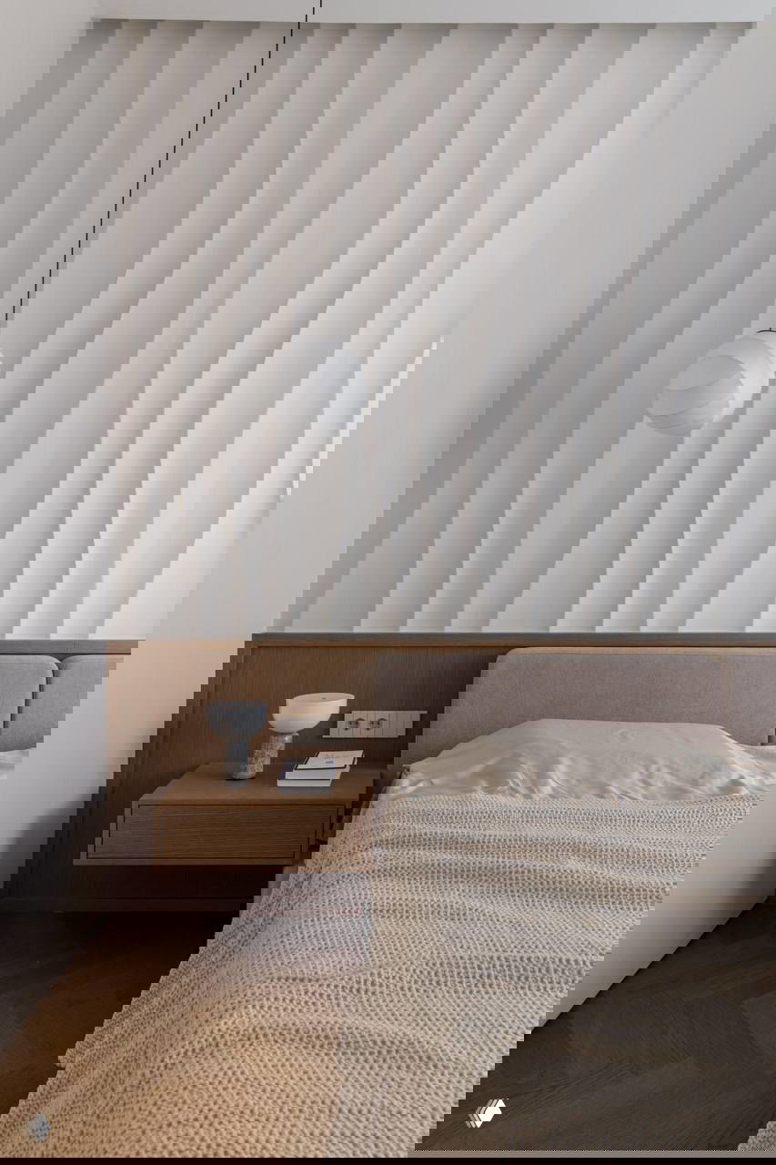 Спальная зона с текстурной волнообразной панелью над изголовьем, тёплая деревянная тумба и спокойная светлая палитра для расслабляющего интерьера.
