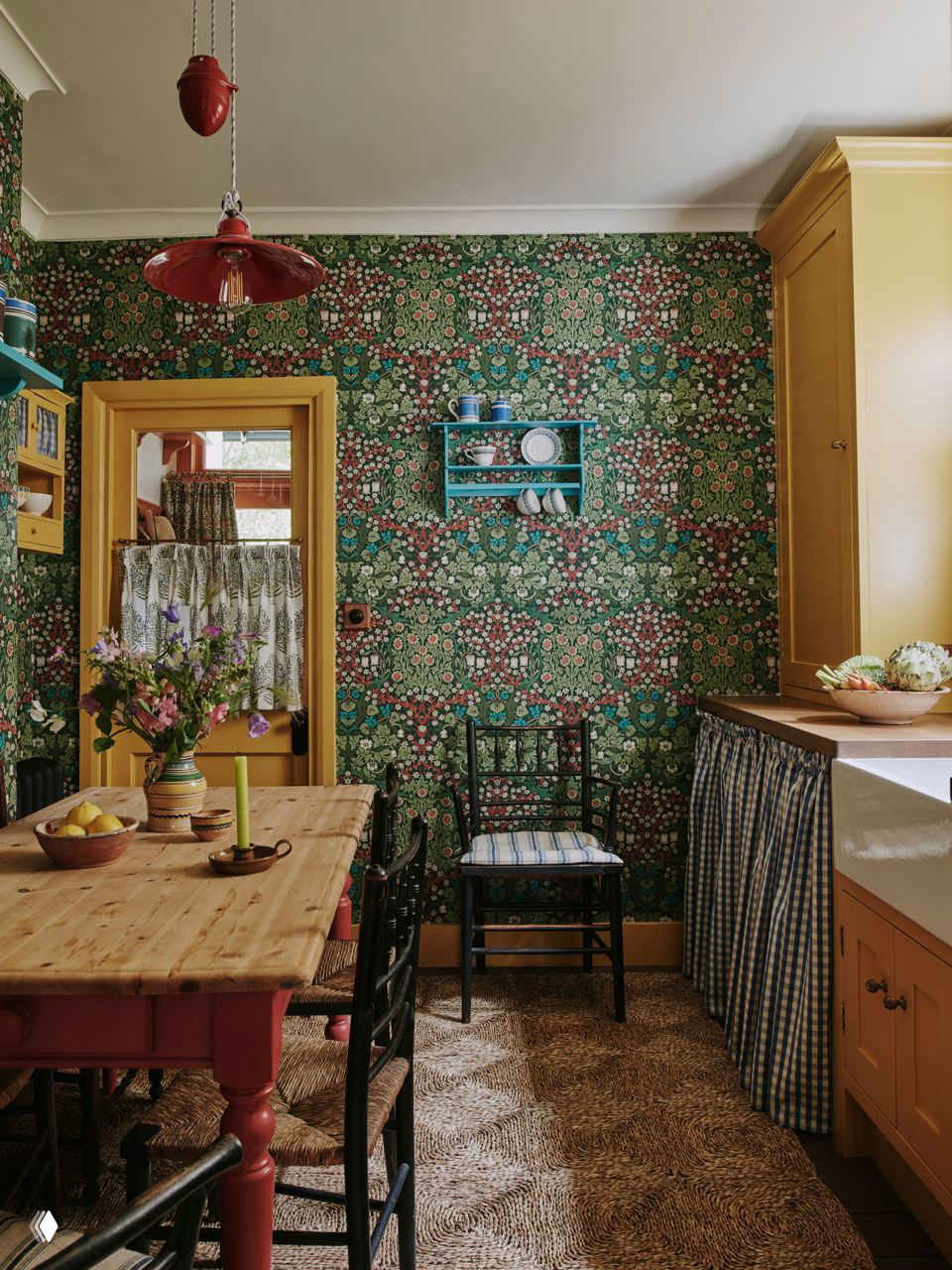 Кухня с насыщенными зелёными обоями в орнаменте, жёлтыми наличниками дверей, деревянным столом и сине-белой шторкой, скрывающей бытовую технику.