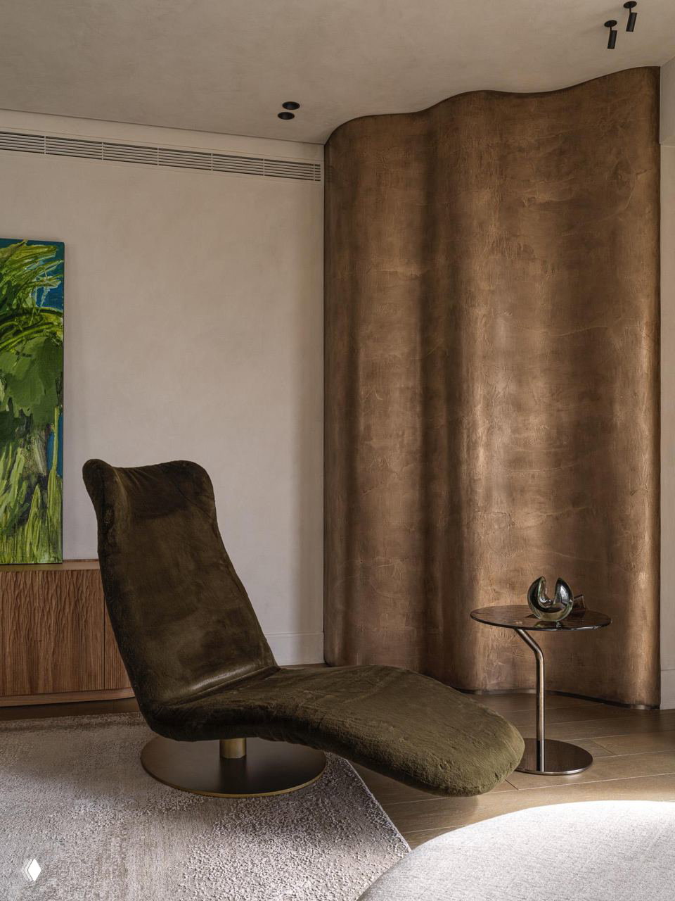 Крупный план шезлонга в оливково-коричневой обивке на фоне изогнутой бронзовой панели и нейтральной стены, акцент на форму и фактуру материалов.