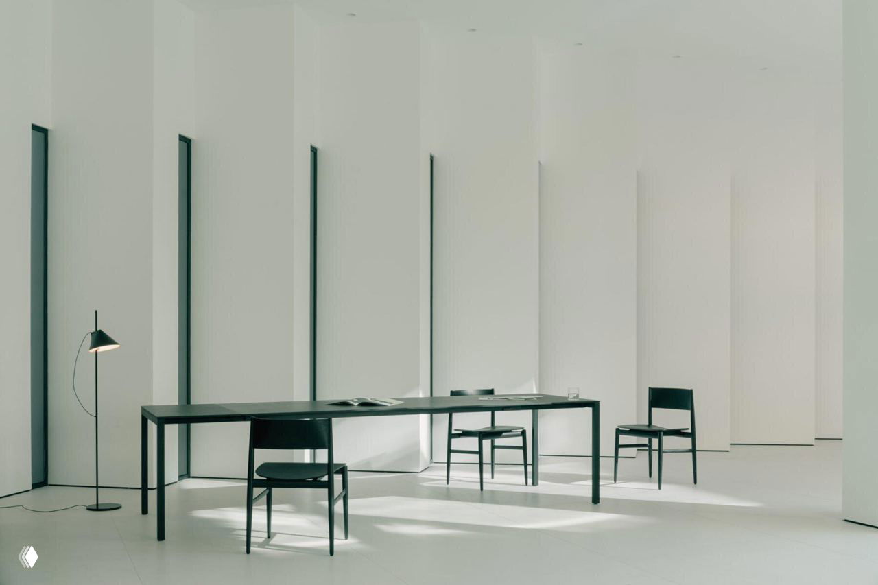 Интерьер: минималистичная светлая комната с высокой потолочной высотой, тонкими вертикальными окнами и лаконичной мебелью на фоне белых стен.