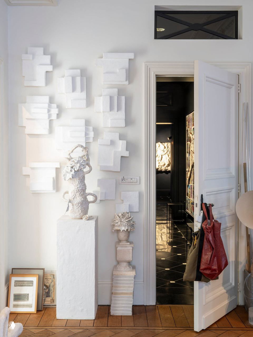 Узкий коридор или входная зона с белыми настенными рельефными панелями и скульптурой на постаменте, деревянный паркет и дверь в темную комнату.