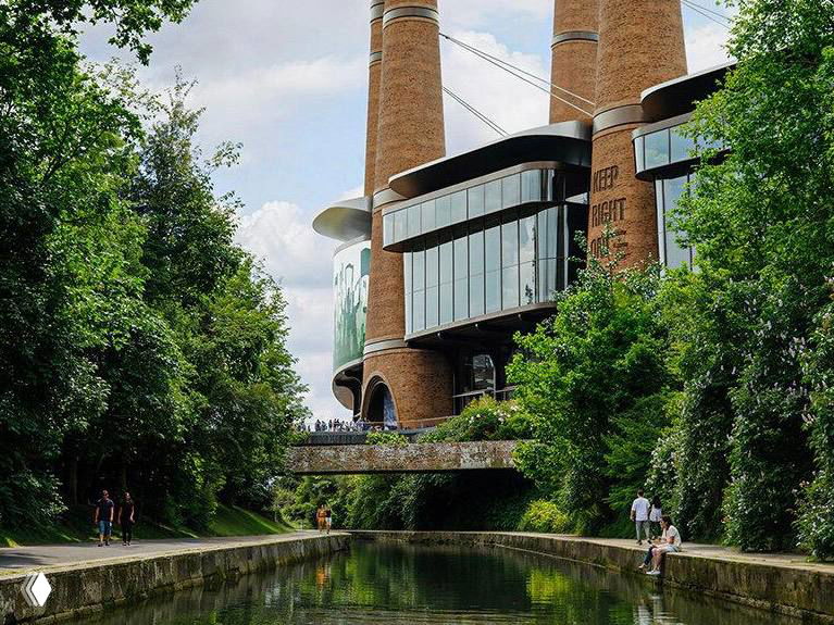 Ближний план одной из башен‑дымоходов с панорамными элементами и входной зоной у воды, подчёркивающий материалы и детали фасада.