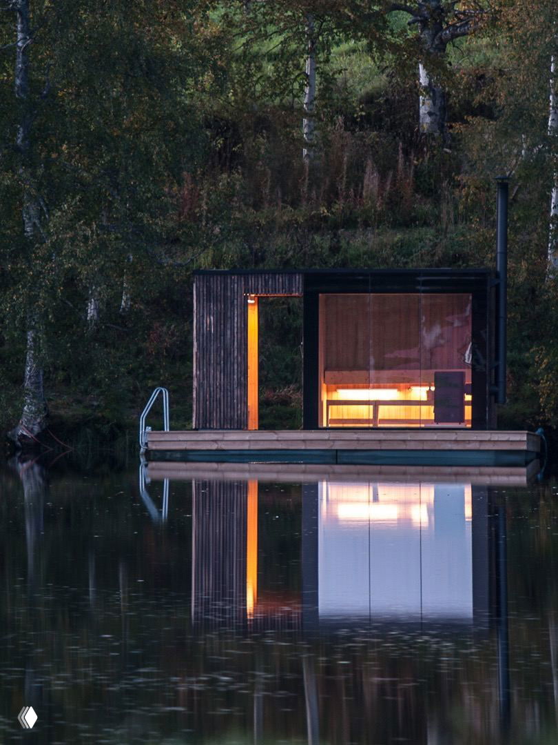 Ночная фотография маленькой бани на плавающей платформе у лесного берега, тёплое внутреннее освещение и отражение в спокойной воде.