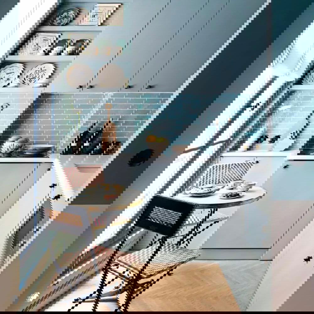 Небольшая кухня в пастельных голубых тонах: фасады, фартук из плитки-кабанчик, окно, небольшой круглый стол и плетёные стулья у окна.